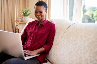 Genç Afrikalı Amerikalı kadın evinde dizüstü bilgisayarla çalışırken oturma odasındaki kanepede dinlenirken gülümsüyor.  