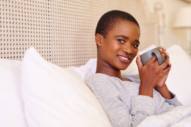 Sabahları yatağında oturmuş bir fincan kahve içen gülümseyen Afro-Amerikan bir kadının portresi.