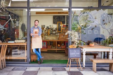 Moda bir kafenin kapısında kollarını kavuşturmuş gülümseyen bir baristanın portresi. 