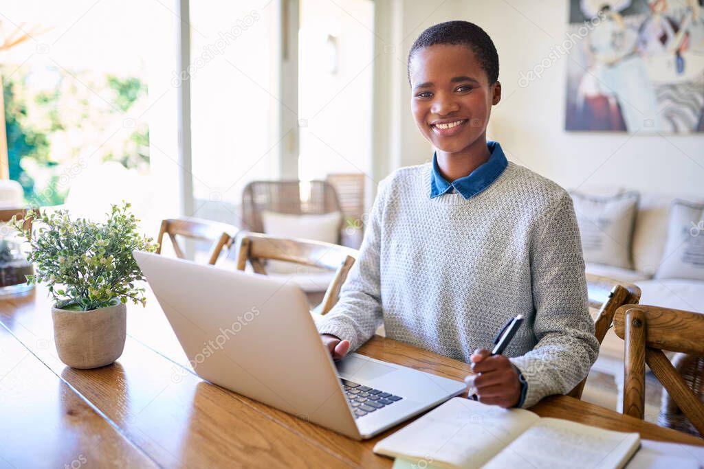 Portrait of a smiling young African American female entrepreneur working with a laptop and taking notes while sitting at her table at home 
