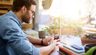 Genç adam kafenin penceresinin yanındaki masada tek başına otururken cep telefonundan mesaj okuyor. 