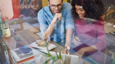Çeşitli genç iş arkadaşları bir kafenin penceresinde otururken birlikte gülümsüyor ve dizüstü bilgisayar kullanıyorlar.