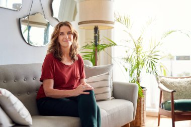 Güneşli evinde kanepede bacak bacak bacak üstüne atmış, gülümseyen olgun bir kadının portresi.