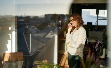 Evinin içinde pencerelerinde şehrin yansımasıyla cep telefonuyla konuşan gülümseyen olgun kadınlar.