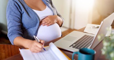 Hamile bir kadının evde yemek masasında doğum izni alırken not yazıp dizüstü bilgisayar kullanması. 