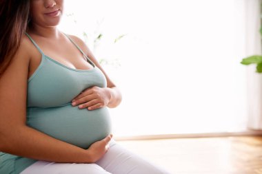 Gülümseyen hamile bir kadın evinde dinlenirken şefkatle karnını sarıyor.