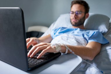 Bir hastane yatağında yatan ve laptopunda çalışan damar içi bir damlaya bağlı bir hastalıktan iyileşen genç adam.  