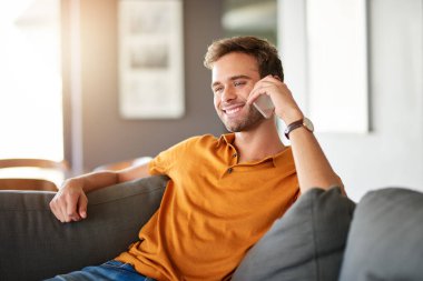 Oturma odasındaki kanepede tek başına oturan gülümseyen genç adam cep telefonuyla bir arkadaşıyla konuşuyor. 