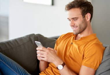 Gülümseyen genç adam oturma odasındaki kanepesinde yalnız başına cep telefonuna mesaj atıyor.  