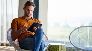 Genç adam modern dairesinde bir sandalyede tek başına oturmuş dijital tabletle internette çalışıyor.