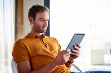 Dairesinde bir sandalyede oturmuş dijital tabletle çalışan gülümseyen genç adam.