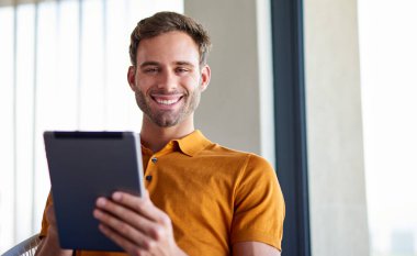 Dairesinde bir sandalyede oturmuş dijital tabletle çalışan gülümseyen genç bir adamın portresi.