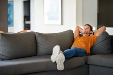 Genç adam oturma odasındaki kanepesinde gözleri kapalı ve elleri başının arkasında dinleniyor. 