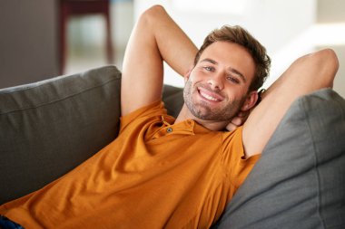 Oturma odasındaki kanepede ellerini başının arkasına koyup rahatlayan gülümseyen genç bir adamın portresi. 