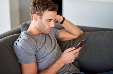 Genç bir adam evdeki kanepede tek başına otururken cep telefonuyla mesajlaşıyor.