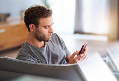 Genç adam evdeki oturma odasındaki kanepede dinlenirken cep telefonundan mesaj okuyor.