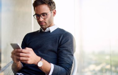 Genç bir girişimci cep telefonuna mesaj yollamak için evdeki sandalyesine yaslanıyor.