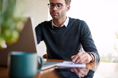 Odaklanmış genç işadamı evde bir masada oturuyor bir dizüstü bilgisayar üzerinde çalışıyor ve bir deftere fikirler yazıyor.