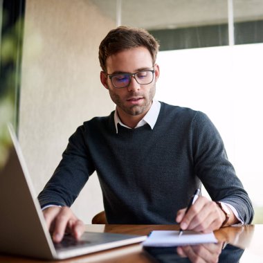 Not defterine fikirler yazan ve evde bir masada otururken dizüstü bilgisayarda çalışan genç bir işadamı. 