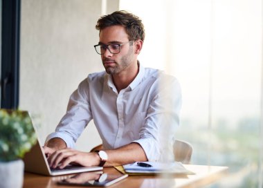Sıradan giyinmiş, gözlüklü genç bir işadamı evde oturmuş dizüstü bilgisayarla çalışıyor.