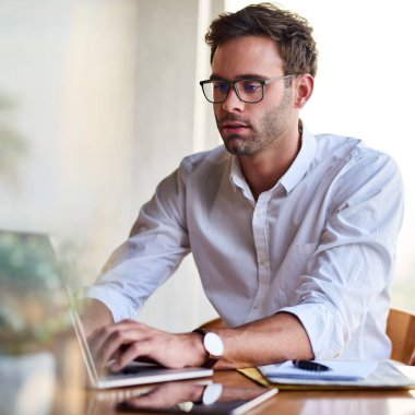 Evde masa başında çalışırken dizüstü bilgisayarda gözlük takan sıradan giyinmiş genç bir iş adamı. 