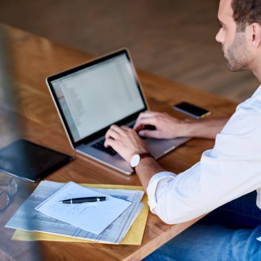 Odaklanmış genç işadamı evde bir masada oturuyor bir dizüstü bilgisayar üzerinde çalışıyor ve notları inceliyor.