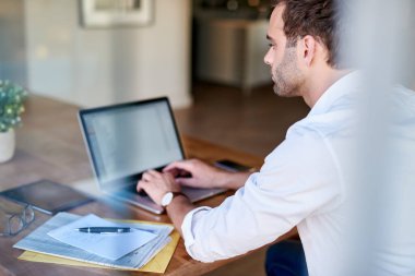 Sıradan giyimli genç iş adamı dizüstü bilgisayarda çalışıyor ve evdeki masada otururken evrakları inceliyor. 