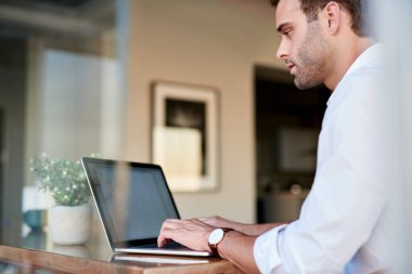 Sıradan giyimli genç iş adamı evde yemek masasında tek başına oturmuş bir dizüstü bilgisayarla çalışıyor.