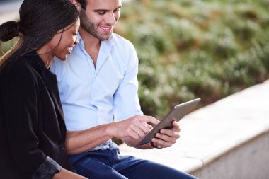 Ofis binasının dışında oturmuş dijital tablet üzerinde çalışan iki farklı iş adamı gülümsüyor.