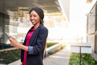 Bir ofis binasının önünde dijital tablet üzerinde çalışan gülümseyen Afrikalı genç bir iş kadınının portresi. 