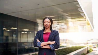 Bir ofis binasının önünde kollarını kavuşturmuş kendinden emin bir Afrikalı Amerikalı iş kadınının portresi.