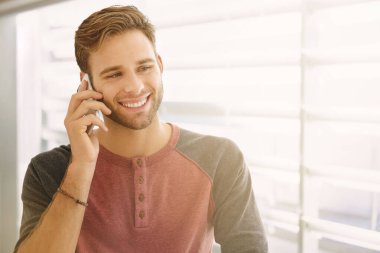 Kapalı alanda telefonunu kullanan yakışıklı genç adam, dinlerken ve bakarken gülümsüyor, güneş ışığıyla.