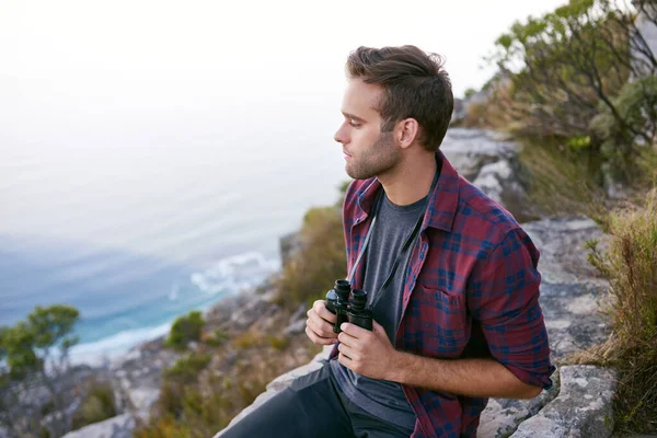 Dürbün tutan yakışıklı bir delikanlının yüksek açılı görüntüsü. Dağdaki kayalık bir çıkıntıda otururken. Altında okyanus görünüyor.