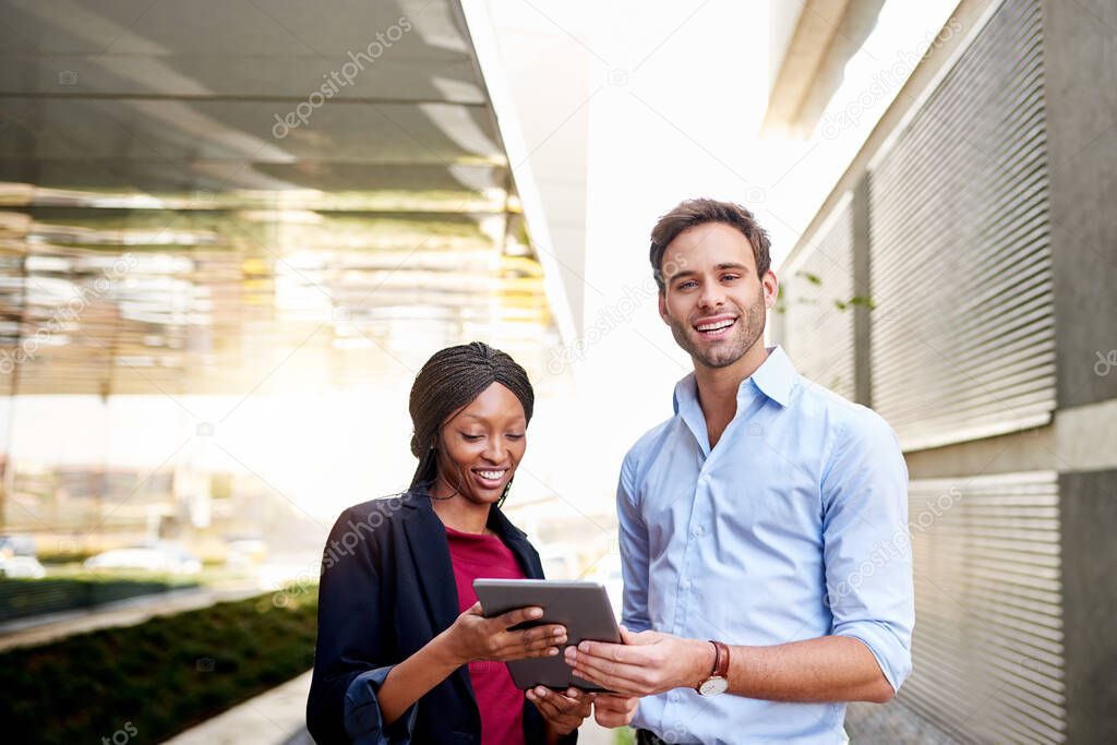 Portrait of two smiling diverse businesspeople standing outside of an office complex working on a digital tablet