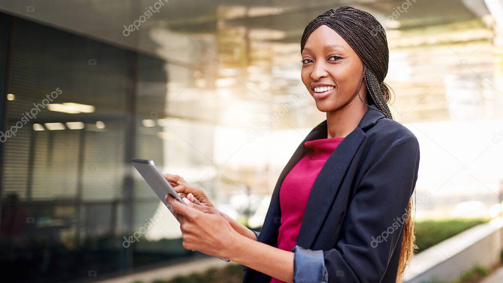 Portrait of a smiling young African American businesswoman standing outside of an office building working on a digital tablet