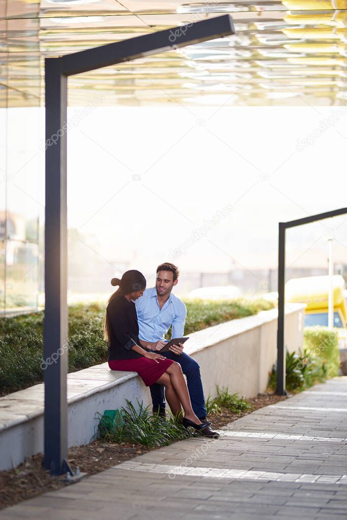 Two diverse young businesspeople sitting outside an office building talking together and working on a tablet