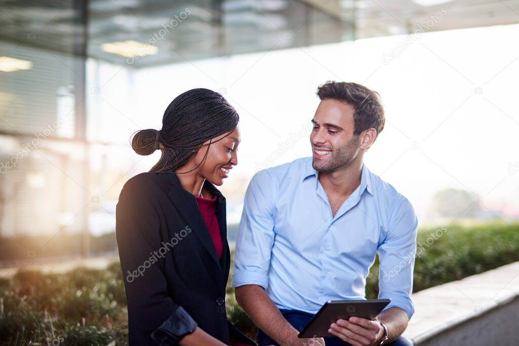 Two young businesspeople laughing while sitting together outside working on a digital tablet
