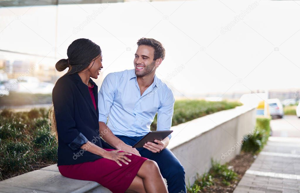 Two laughing young businesspeople sitting together outside their office building working on a digital tablet