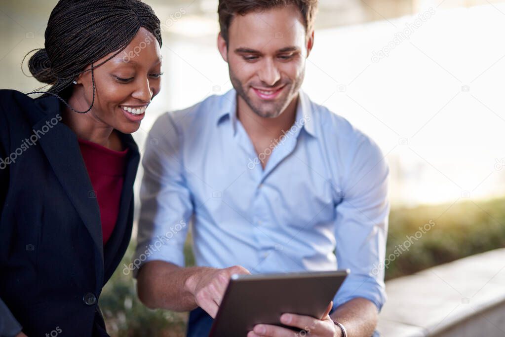 Two diverse young businesspeople sitting together outside and smiling while discussing a project on a digital tablet 