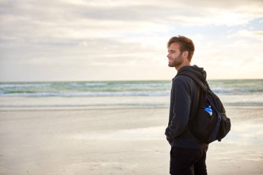 Genç adam sabahın köründe bir kumsalda tek başına kapüşonlu ve sırt çantalı dikilirken düşünceli görünüyor.