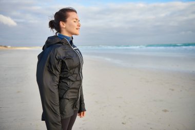 Gözleri kapalı bir kadın, kışın kumsalda temiz deniz havasını solurken pozitifliğini ifade ediyor.