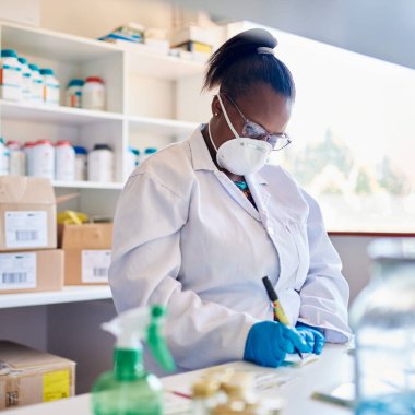 Palto giymiş Afrikalı kadın teknisyen ve koruyucu yüz maskesi. Laboratuvardaki bir masada çalışırken verileri yazıyor.