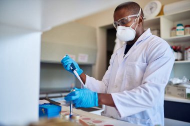 Koruyucu maskeli ve gözlüklü Afrikalı erkek teknisyen laboratuarda çalışırken örnek hazırlamak için pipet kullanıyor.