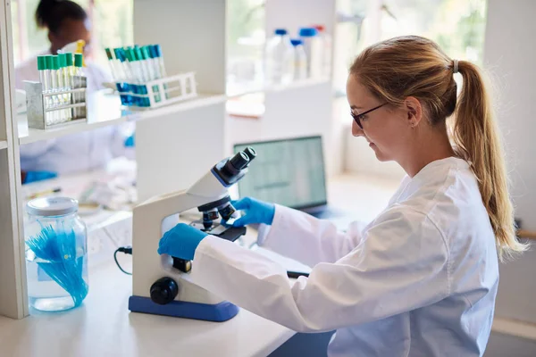 Female lab technician placing a slide with a sample under a microscope while sitting at a table in a lab