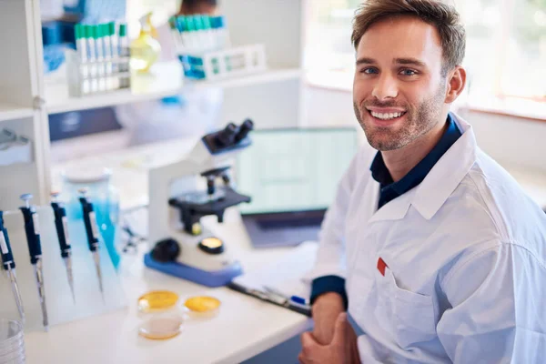 Portrait of a smiling young male lab technician sitting at a table analyzing samples with a microscope