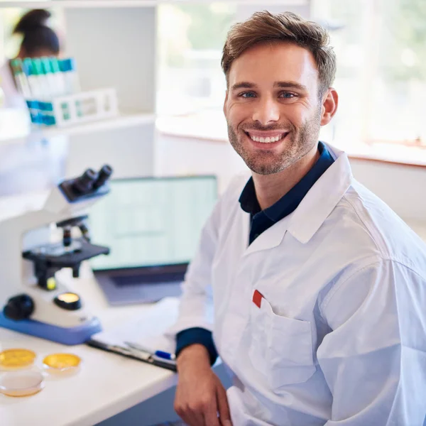 Portrait of a young male lab technician smiling while sitting at a table analyzing samples with a microscope