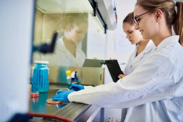 Female lab technicians analyzing a sample in a petri dish inside of a biosafety cabinet