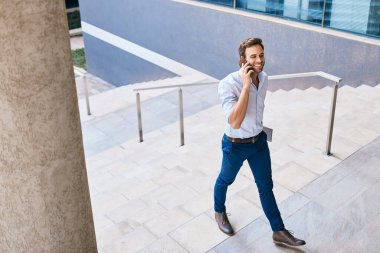 Modern bir ofis binasının önünde merdivenlerden çıkarken gülümseyen genç bir işadamı cep telefonuyla konuşuyor.