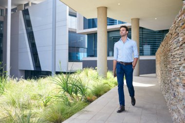 Modern bir ofis binasının önündeki patikada yürüyen dijital tabletli kendinden emin genç bir işadamı.