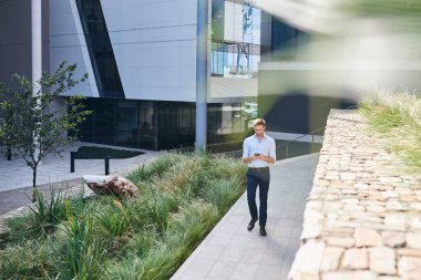 Genç bir işadamı modern bir ofis binasının önündeki patikada yürüyor ve cep telefonuyla mesajlaşıyor.